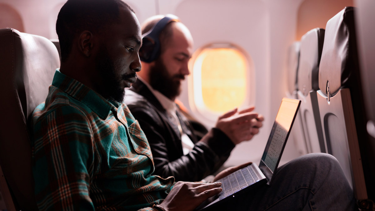two people on a plane, one wearing heaphones, the other looking at a laptop