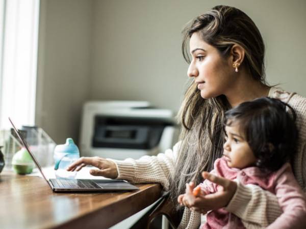 Mother holding baby in one arm while typing on the laptop with other hand