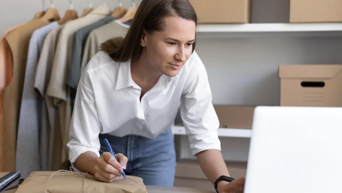 A woman in a white shirt and jeans is leaning over a table and writing on a package with a pen. In the background, there are clothes hanging on a rack and cardboard boxes on shelves. 